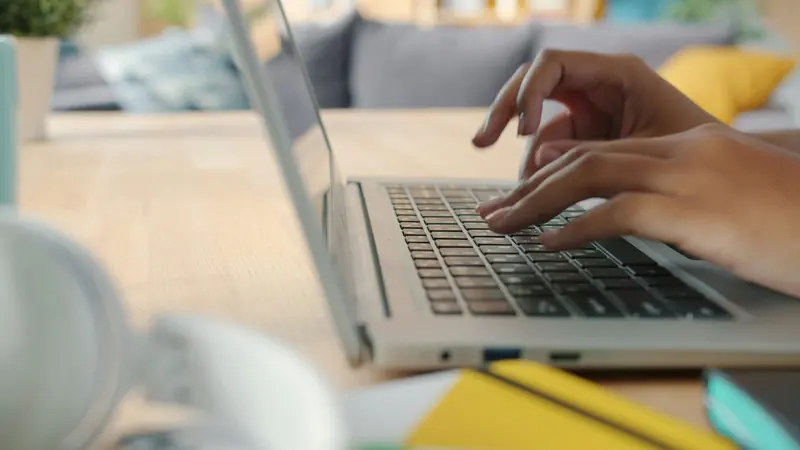 Hands typing on an unbranded silver laptop on a light wooden table with a yellow notebook in the foreground