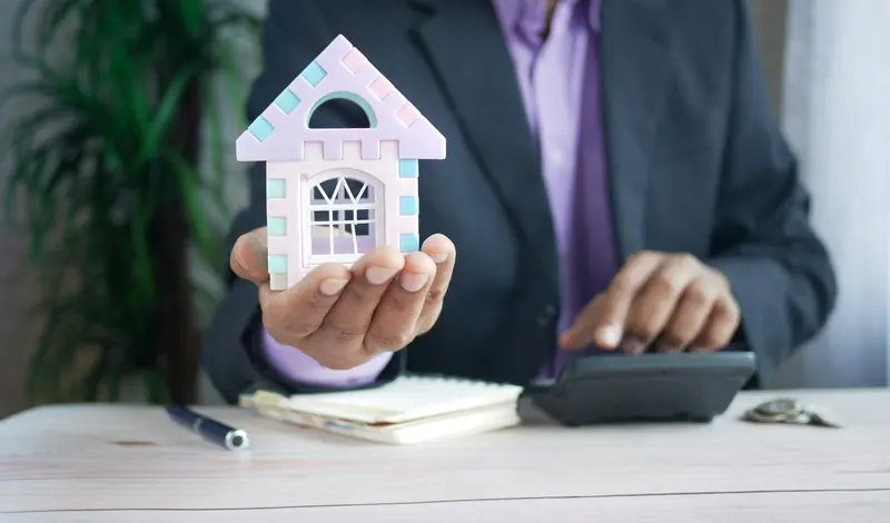 A hand in a dark suit sleeve holds a small pastel model house above a wooden desk with a calculator, notebook and coins