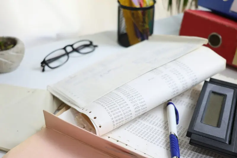 A desk with printed ledger papers, black glasses, a calculator, a pen and binders
