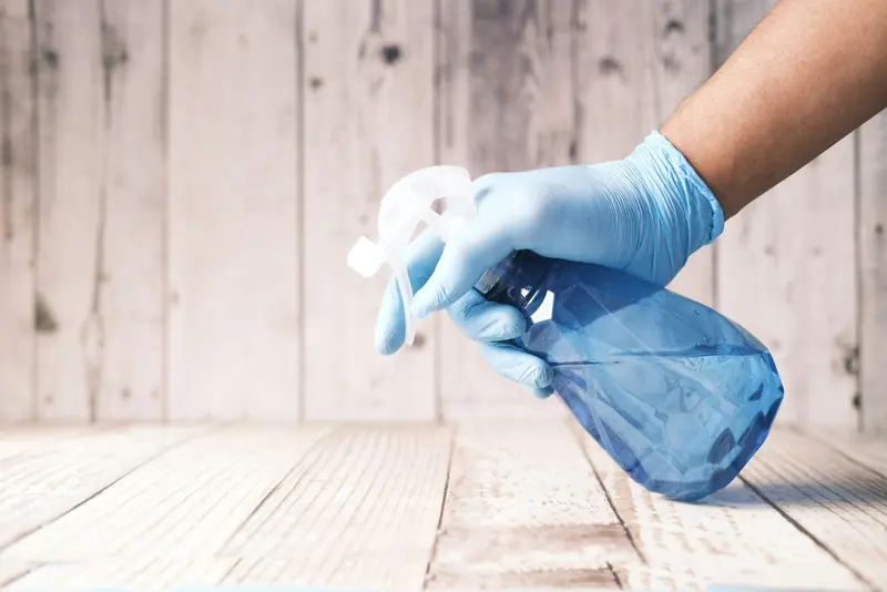 A hand in a blue glove holding a blue spray bottle against a pale wooden background