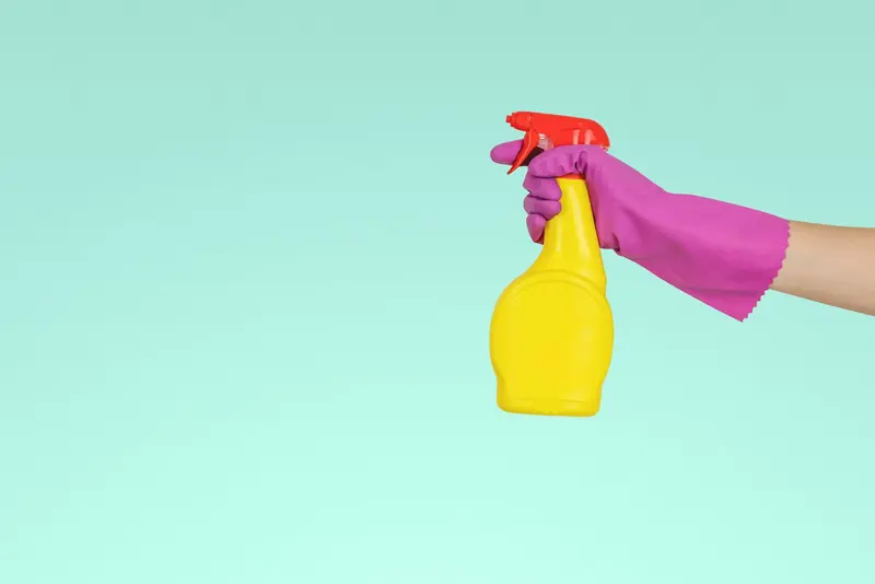A hand in a pink rubber glove holding a yellow spray bottle against a mint green background