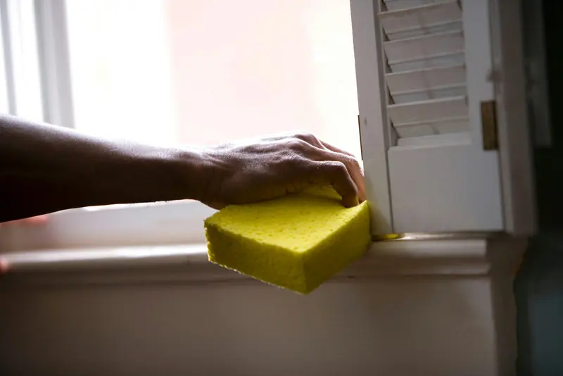 A hand pressing a yellow sponge against a white window sill beside a louvered shutter
