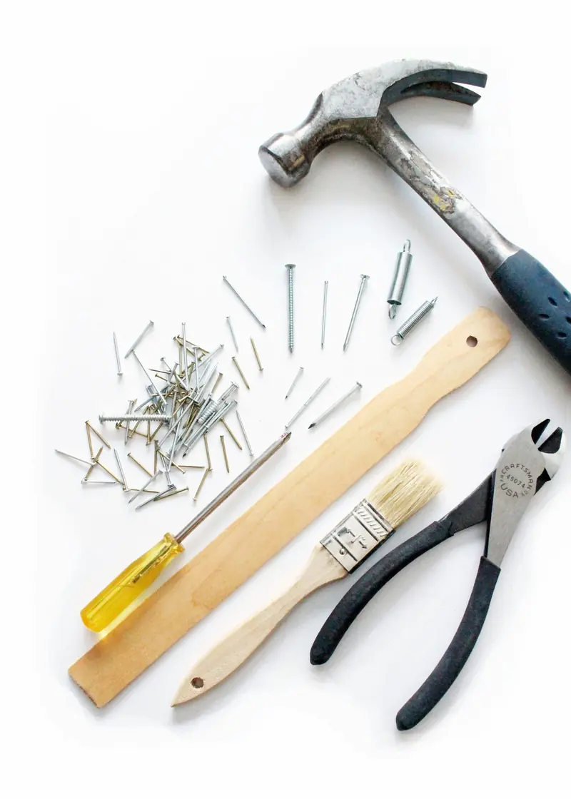 A hammer, nails, screwdriver, wooden ruler, paintbrush and pliers laid out on a white background