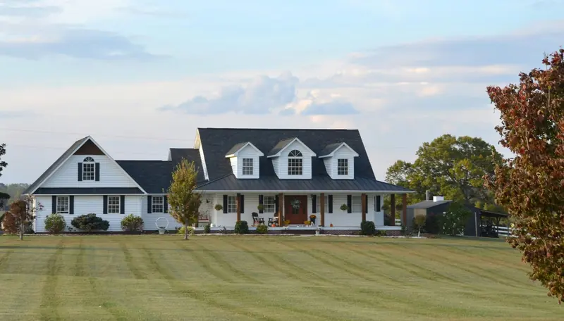 A white farmhouse with dark shutters, dormer windows and a wide front porch behind a freshly mowed lawn